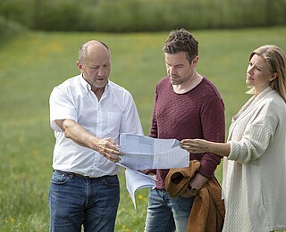 Bild mit drei Menschen die auf einer Wiese stehen und einen Bauplan in der Hand halten